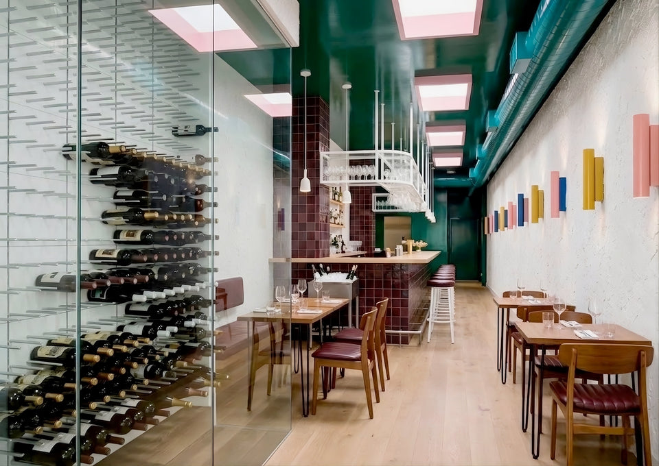 A modern restaurant interior with wooden dining tables and chairs, a glass wine rack filled with bottles on the left, a dark green ceiling, and colorful wall-mounted light fixtures on the right.