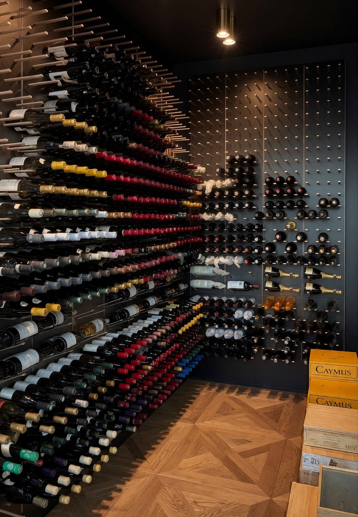 Large custom wine cellar with hundreds of bottles stored horizontally across a floor-to-ceiling wall, a black modular wine display at the rear, warm ceiling spotlights, and geometric wood flooring.