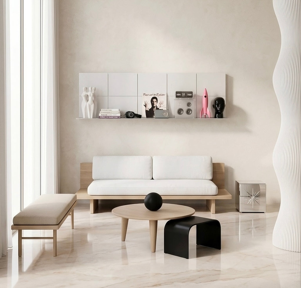 A modern living room scene featuring a white two-seater sofa with a light wood frame, a matching beige bench, a round light wood coffee table, and a black sculptural side table. The room has a warm beige wall with a floating shelf holding decorative items, and a large white ribbed column on the right.