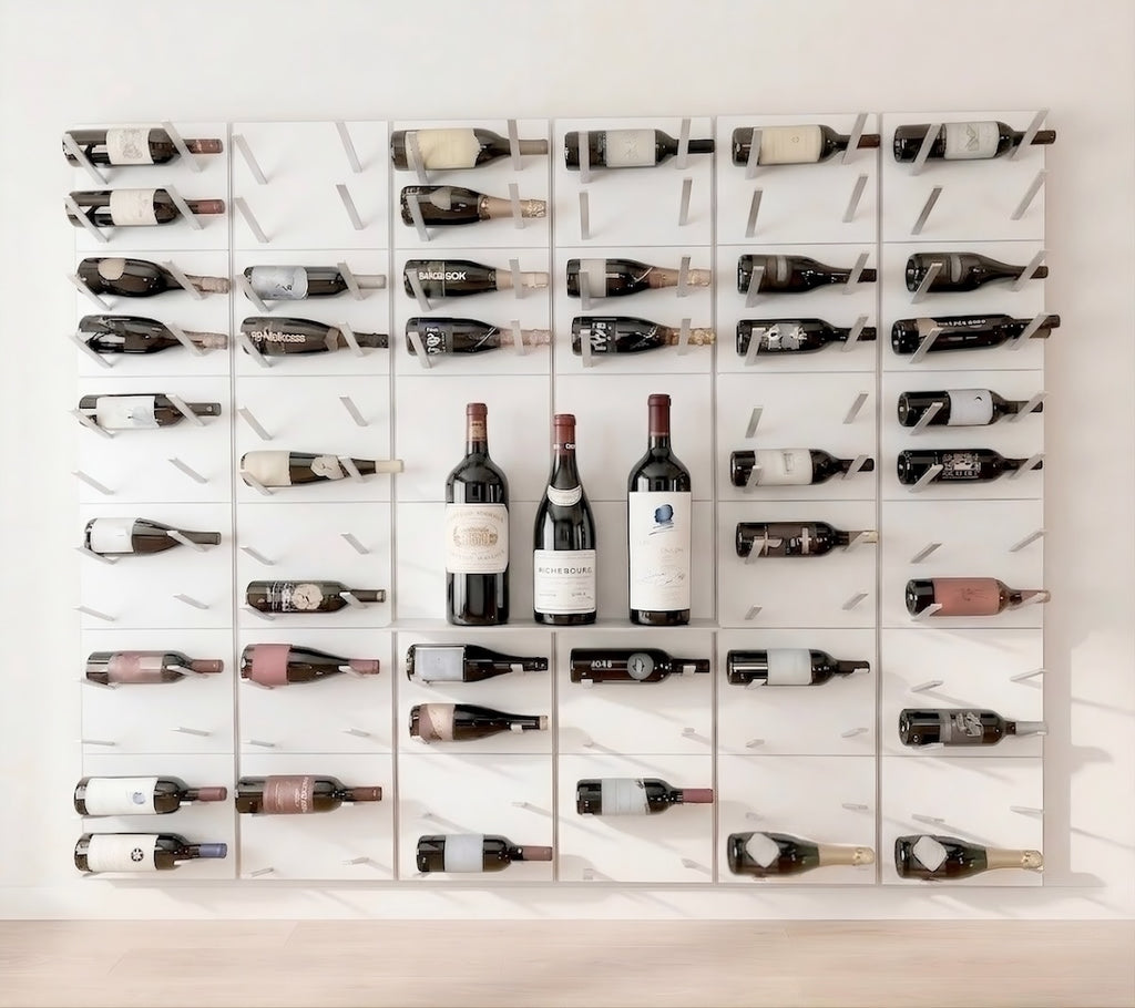 A wall-mounted wine rack made of white rectangular panels with sleek angled silver metal bottle support bars, and a cantilevered display shelf, holding multiple wine bottles arranged in a grid.
