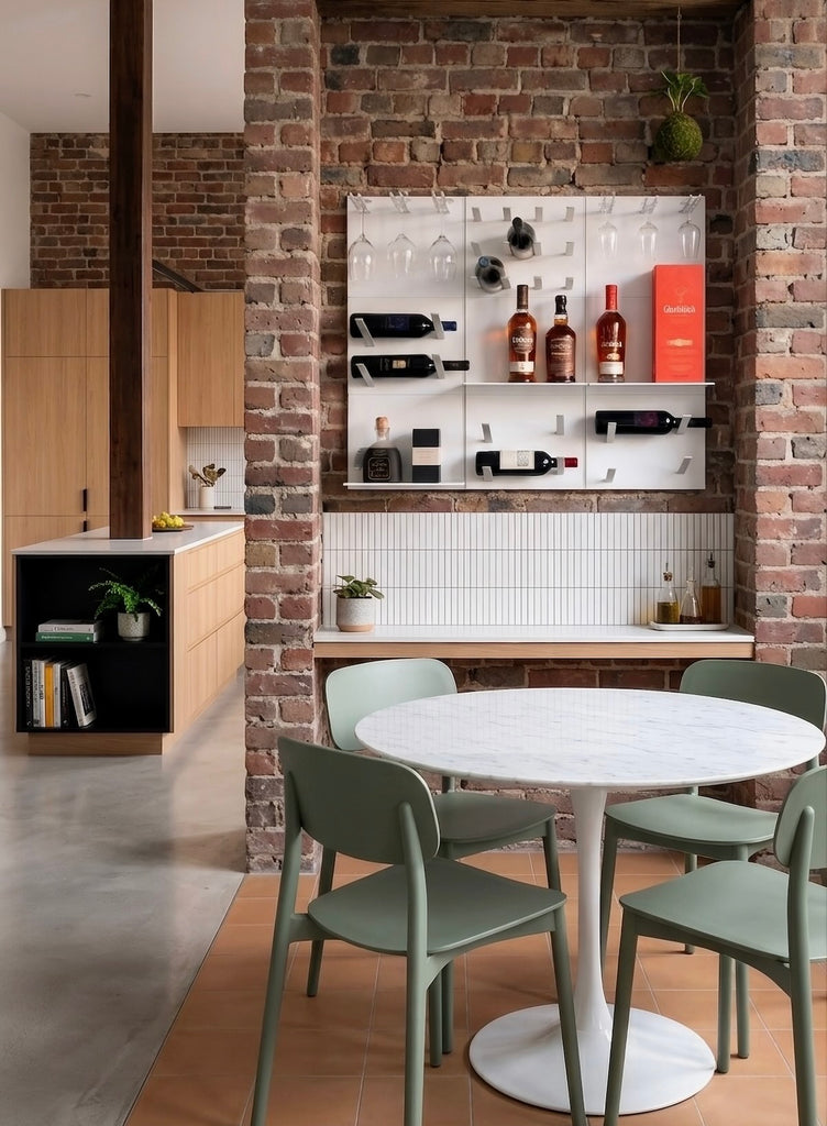 A modern dining area with a round white marble-top table and four sage green chairs, set against a brick wall with a white wine rack and bar counter.