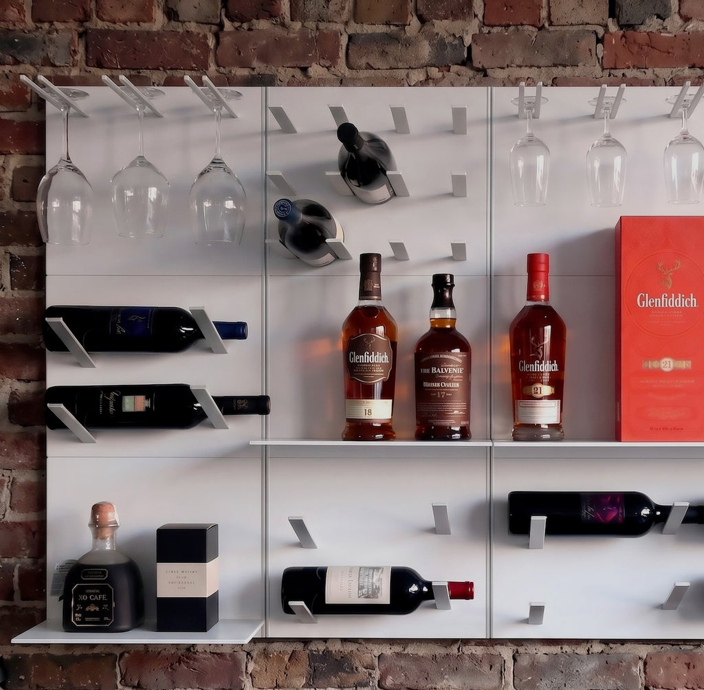 A white wall-mounted wine rack with multiple shelves and glass holders, styled with several wine bottles, wine glasses, and a red Glenfiddich box against a rustic brick wall.